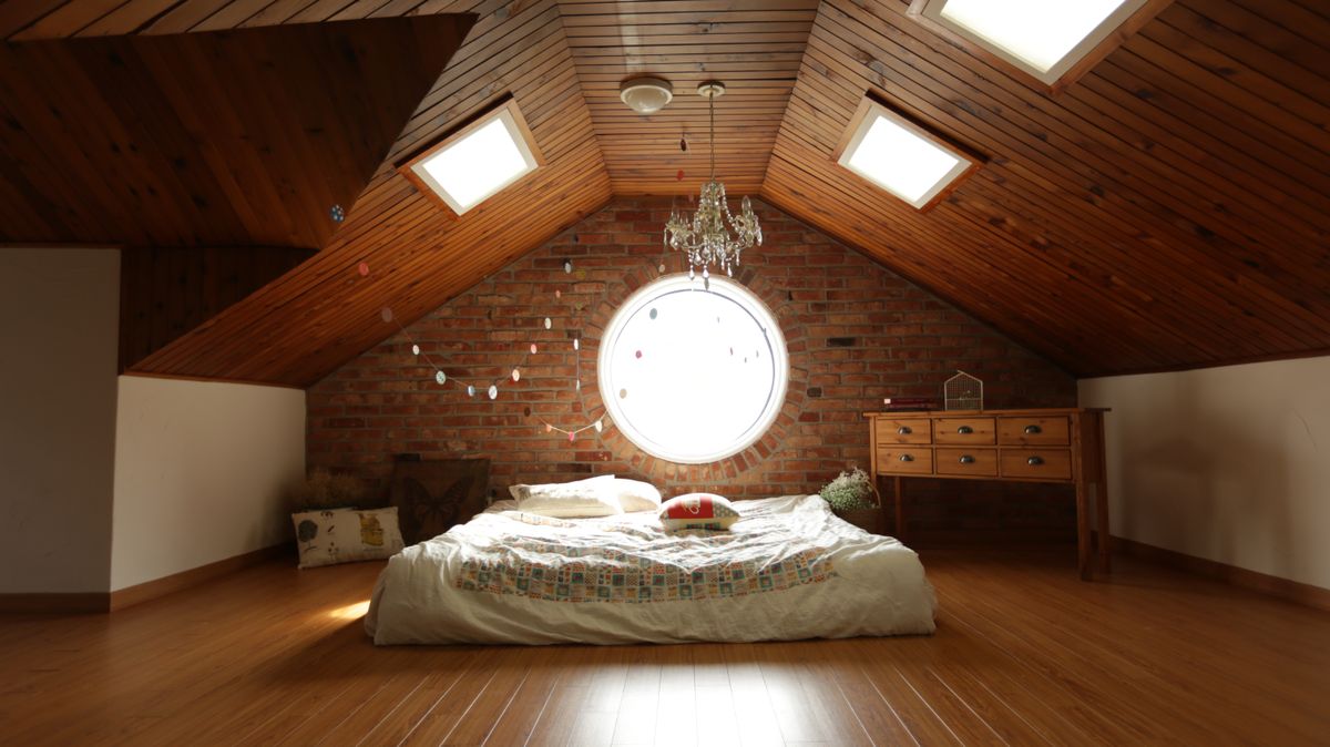 Attic bedroom with sloped wooden ceiling, skylights, brick wall with circular window, and mattress on a polished wood floor