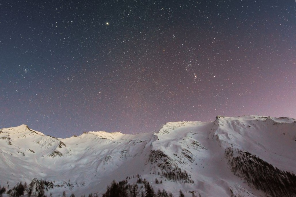 Snow-covered mountain peaks with silhouetted evergreen trees under a star-filled night sky